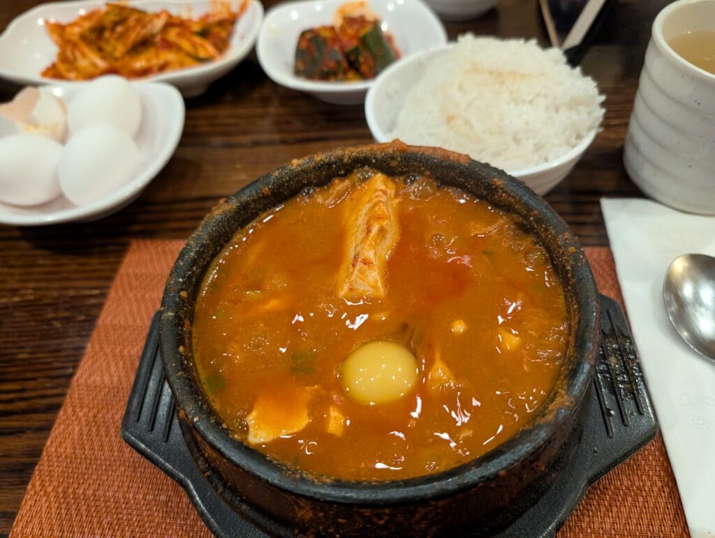 A bubbling stone pot of spicy red Soondubu Jjigae (soft tofu stew) with a raw egg cracked into the center, surrounded by side dishes like kimchi.