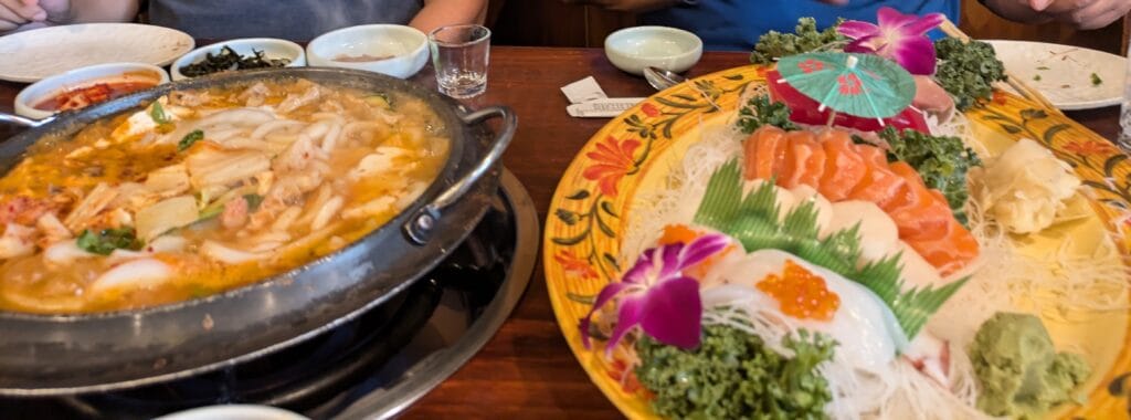 Table spread at Yechon Korean Restaurant with a simmering seafood jonggol hot pot and a vibrant sashimi platter featuring salmon, scallops, and squid.