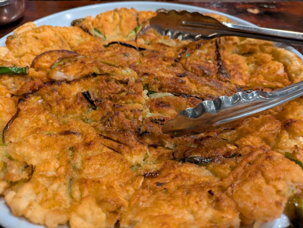 Crispy, golden-brown Haemul Pajeon (Korean seafood and green onion pancake) served with tongs at Yechon Korean Restaurant.