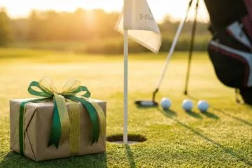A wrapped gift box with green and gold ribbon sitting on a sunny golf course green next to the flagstick and hole, with a golf bag in the background.